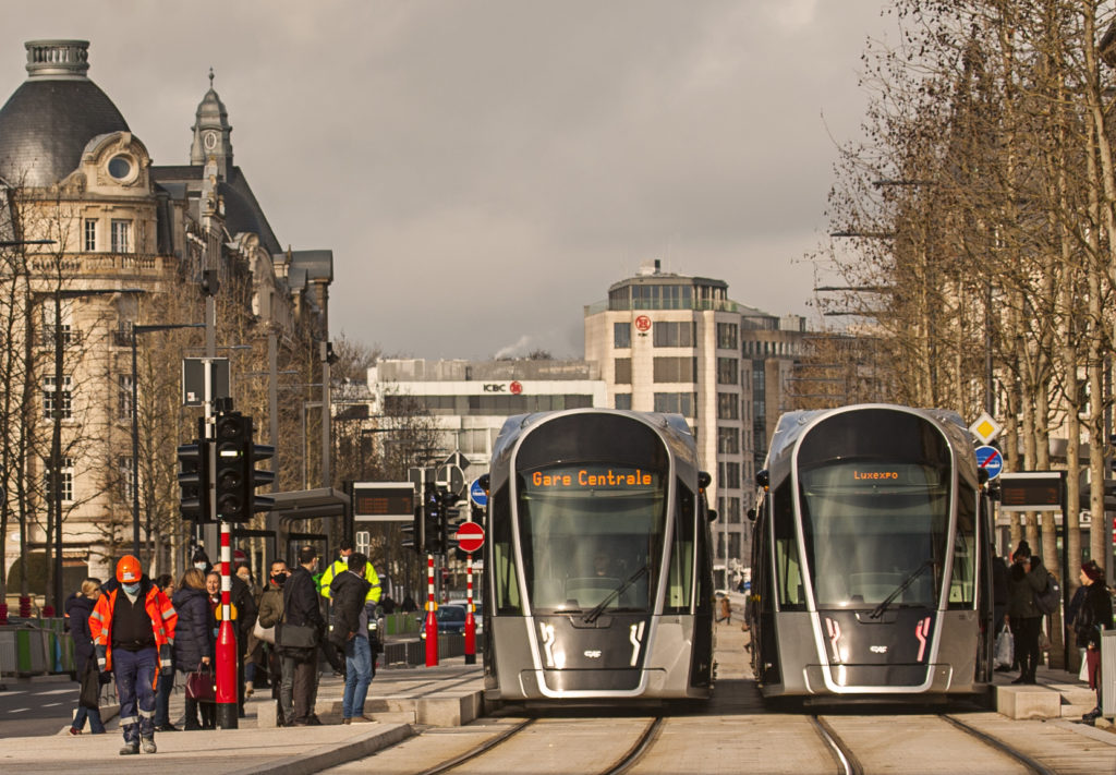 en la avenida de la libertad se encuentran en una parada del tram se encuentran los dos trenes con las personas en tránsito para salir y entrar o abordar y continuar la vía que atravieza la ciudad de Luxemburgo